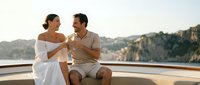 Romantic couple toasting with champagne glasses on luxury yacht deck during sunset cruise with scenic coastal cliffs in background for anniversary celebration.