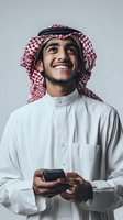 Happy young Middle Eastern businessman in traditional white thobe and red checkered keffiyeh holding smartphone, smiling while looking up on gray background.