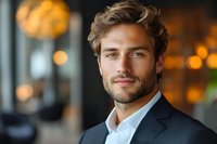 Confident young Caucasian businessman with beard in dark suit smiling at camera in modern office with warm bokeh lights background for corporate marketing.