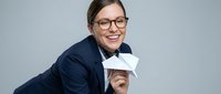 Young woman with dark hair, glasses, and fair complexion wearing navy blazer holds blank white card against neutral background, suitable for business, corporate messaging, and promotional campaigns.