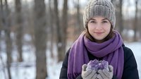 Young woman in knit hat and purple scarf holding snow in cupped hands during winter day outdoors with forest background for seasonal lifestyle content.