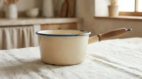 Vintage cream enamel saucepan with wooden handle on linen tablecloth in rustic farmhouse kitchen setting for cooking and food preparation concepts.