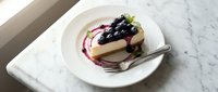 Creamy cheesecake slice topped with fresh blueberries and berry sauce on white plate, elegant dessert presentation for cafes and restaurants.