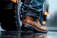 Brown leather boot with rugged sole and detailed stitching on wet asphalt surface, paired with blue denim jeans, captured in moody atmospheric lighting.