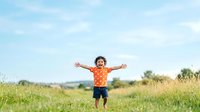 Young boy with curly dark hair and light skin tone wearing orange shirt standing in green meadow with arms outstretched, expressing joy and freedom outdoors.