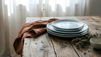Stacked ceramic plates with rust-colored linen cloth on rustic wooden table near window with city skyline view. Styled tableware setup for dining, home decor, and lifestyle photography.
