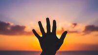 Hand silhouette reaching toward vibrant sunset sky with orange and purple clouds over ocean horizon for inspirational and motivational concepts.