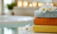 Folded spa towels in orange, yellow and mint colors decorated with white orchid flowers on blurred bathroom background with glowing candles and water.
