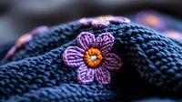 Delicate purple crochet flower with orange center detail on dark blue knitted textile background, showcasing intricate handmade needlework pattern and texture.