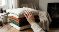 Woman hand touching stack of cozy knitted sweaters on wooden table in warm living room interior for autumn winter fashion wardrobe organization.