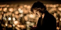 Young man with curly hair using smartphone at night with warm bokeh city lights background for social media and technology concepts.