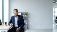 Middle-aged man of South Asian descent wearing navy blazer and light blue shirt, sitting at modern office desk with computer, potted plant and shelving visible in bright contemporary workspace.