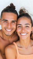 Close up portrait of young Hispanic couple with bright white smiles and tanned skin wearing summer attire, looking at camera against light background.