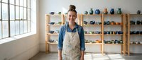Young female potter standing in ceramic studio workshop with handmade pottery displayed on wooden shelves, artisan craftswoman in apron smiling at camera.