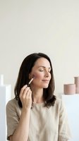 Young woman applying makeup with brush in modern bathroom setting with cosmetic products on counter for beauty routine and skincare lifestyle.