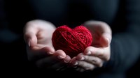 Red yarn heart shape held gently in cupped hands against dark background, symbolizing care, love, and tenderness. Romantic concept for Valentine's Day and relationships.