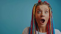 Young woman with colorful braids and shocked facial expression against blue background showing surprise and amazement with wide open mouth and eyes.