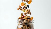Mixed nuts and dried fruits falling into glass jar on white background, ideal for healthy snacking, nutrition, and food storage concepts.