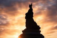 Dramatic silhouette of a classical statue monument against vibrant orange and golden sunset sky with dramatic clouds creating moody atmospheric scene.