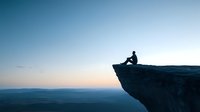 Silhouette of person sitting on cliff edge overlooking misty mountain landscape at dawn, contemplative mood captures solitude and achievement in nature.