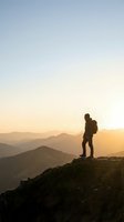 Silhouetted hiker with backpack standing on mountain peak at golden hour sunrise, overlooking layered mountain ranges and valleys below in warm atmospheric light.