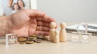 Wooden family figures with protective hand gesture over coins and insurance blocks representing financial security and family protection planning.