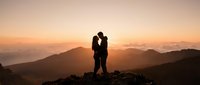 Romantic couple silhouette embracing on mountain peak during golden sunset with layered hills in background for wedding and love concepts.
