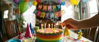 Caucasian hand lighting candles on a colorful rainbow birthday cake at a festive party table with balloons and happy birthday banner.
