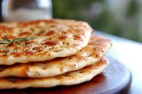 Fresh baked naan bread stacked on wooden board, garnished with herbs and spices, showing golden brown surface with characteristic bubbles and charred spots.