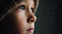 Close-up profile of child with blue eyes looking through rainy window, creating atmospheric mood for concepts of childhood wonder, contemplation, and rainy day emotions.