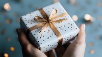 Hands holding white gift box with gold ribbon and polka dot pattern against blurred bokeh background. Festive present wrapped for special occasion or holiday celebration.