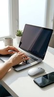 Hands typing on laptop computer at modern workspace with coffee cup, plant and smartphone on white desk near window for remote work productivity.