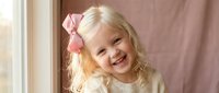 Happy blonde toddler girl with pink bow smiling joyfully in cozy indoor setting with warm natural lighting for family lifestyle photography.