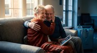 Happy senior Caucasian couple embracing on living room sofa, sharing joyful moment together at home during golden hour sunlight streaming through windows.