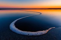 Curved white salt formation along shoreline creates dramatic leading line against vibrant orange and blue sunset sky reflected in still water surface at dusk.