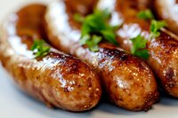 Grilled sausages with golden-brown crust garnished with fresh parsley on white plate, close-up view showing juicy texture and caramelized surface.