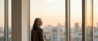 Professional businesswoman in suit looking out office window at city skyline during golden hour sunset, contemplating future opportunities and success.