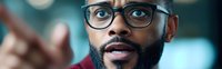 Close up portrait of African American male professional with glasses and beard expressing surprise and pointing forward against blurred background.