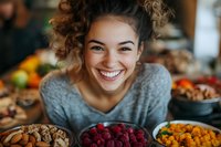 Happy young mixed-race woman smiling at camera surrounded by colorful fresh berries and nuts in cozy kitchen setting for healthy lifestyle content.