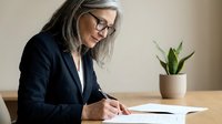 Mature businesswoman with gray hair and glasses writing documents at office desk with plant in background for professional workplace concepts.