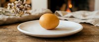Fresh brown chicken egg on white ceramic plate placed on rustic wooden table with cozy fireplace background creating warm homey kitchen atmosphere.