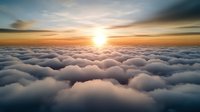 Dramatic aerial view above fluffy white clouds at sunset with golden sunburst on horizon, creating ethereal cloudscape with soft orange and blue sky.