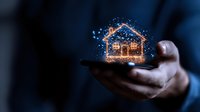 Glowing hologram house floating above smartphone in male hand with sparkles and magical light effects against dark background. Digital real estate concept.