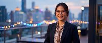 Professional Asian businesswoman in suit smiling confidently against urban city skyline at dusk with illuminated buildings and bridge background.