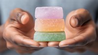 Stack of colorful handmade soap bars in pastel pink, orange and mint green colors held in African American hands against blurred background.