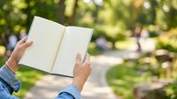 Person with light skin tone holding open blank notebook outdoors in park setting with blurred green foliage background, suitable for education, reading, writing, journaling and lifestyle concepts.