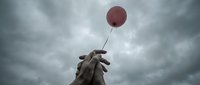 Weathered stone hand sculpture reaching upward holding red balloon against dramatic cloudy sky, symbolizing hope and aspiration for creative projects.