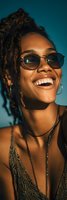 Happy young African American woman with dreadlocks wearing sunglasses laughing joyfully against blue sky background in warm golden hour light.