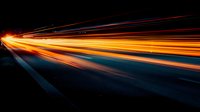Long exposure light trails on dark highway create dynamic motion effect with vibrant orange and blue streaks against black background, showing speed and movement.