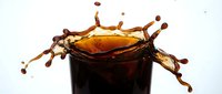 Dark cola beverage splashing in glass with dynamic liquid motion and droplets against white background for refreshment advertising.